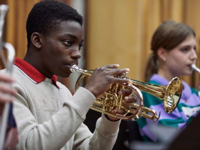 A boy wearing a white jumper, with a red collar sticking out of it, plays the trumpet